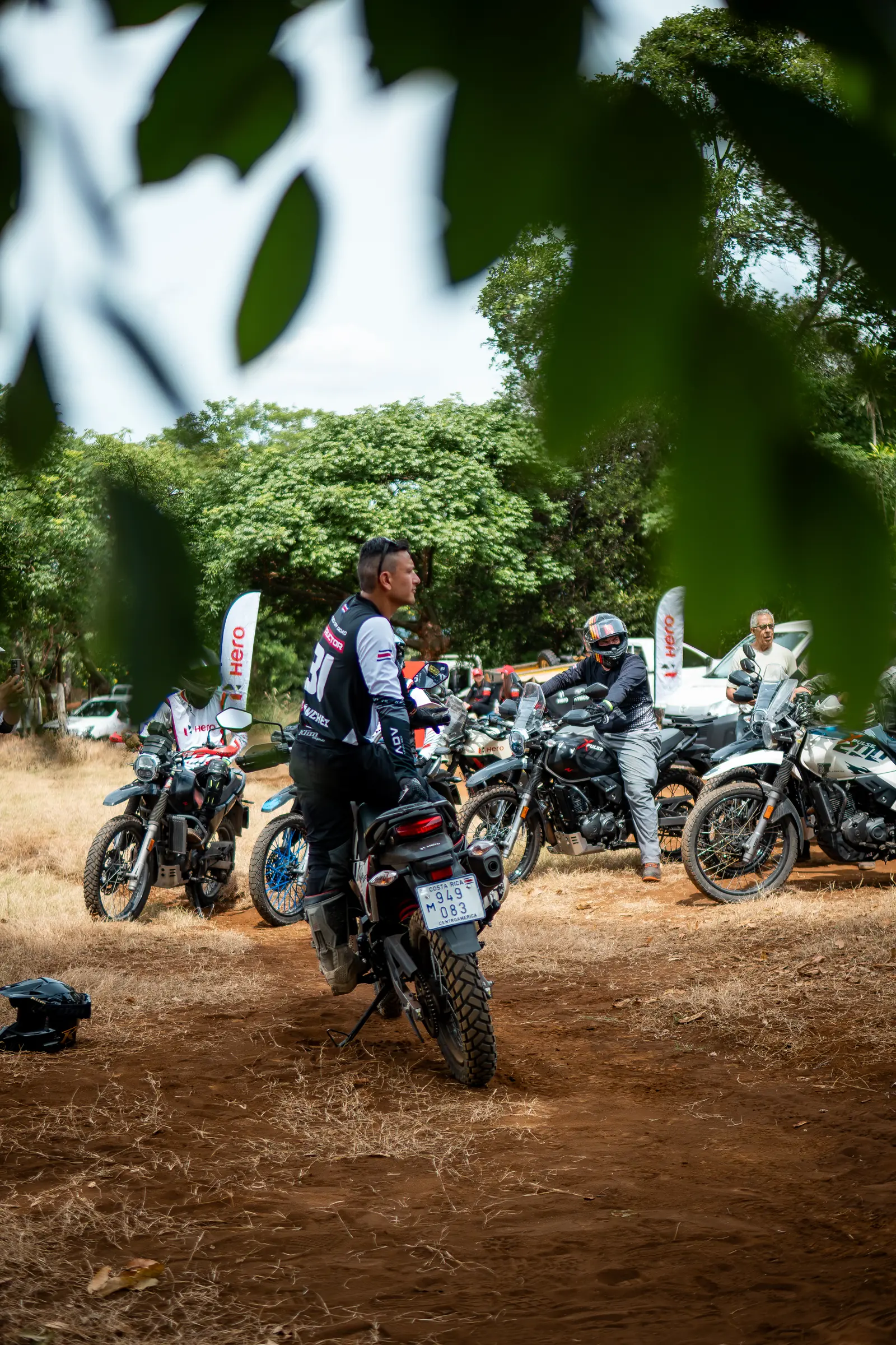 Group of motorcycles and riders gathered under trees at an event